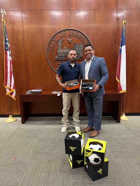 Andrew Zamarripas and a City of Houston official holding donated cleats, with boxed footballs in front of the city seal